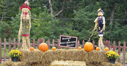 Two scarecrows stand on a hay bale display with pumpkins and flowers, next to a sign for Riverside & Great Northern Railway.
