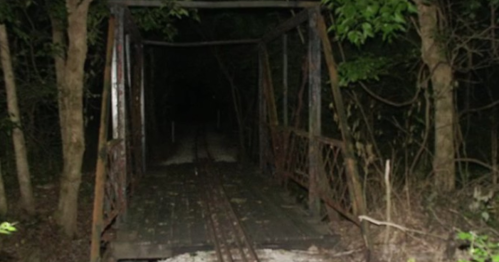 A dark, abandoned bridge surrounded by trees, with faint tracks visible on the wooden surface.