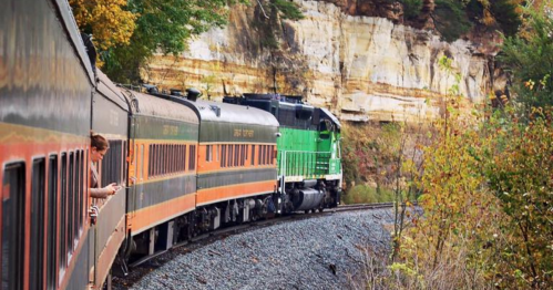 A green locomotive pulls vintage passenger cars around a curve, surrounded by colorful autumn foliage and rocky cliffs.