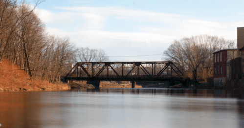 A tranquil river scene featuring a metal bridge and bare trees along the banks under a clear blue sky.