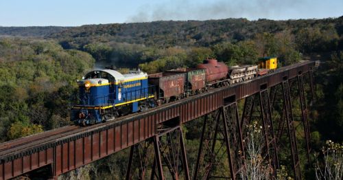 A blue and yellow train crosses a tall bridge, surrounded by lush green trees and a smoky background.