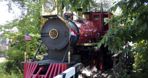A vintage steam locomotive with a shiny black and red exterior, surrounded by lush greenery and American flags.