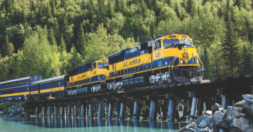 Two yellow and blue Alaska Railroad trains cross a wooden bridge surrounded by lush green trees and a calm body of water.