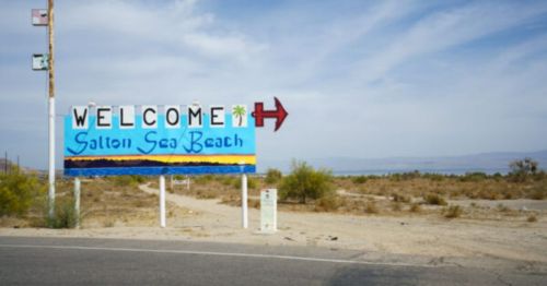 A colorful welcome sign for Salton Sea Beach, with a palm tree and an arrow pointing right, set against a desert landscape.