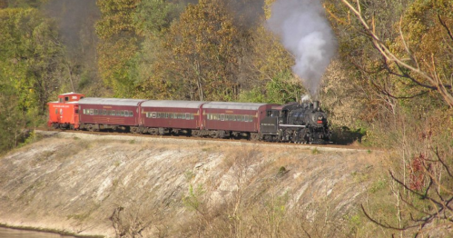 A vintage steam train with a red caboose travels along a scenic route surrounded by autumn foliage.