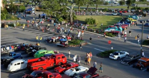 Aerial view of a lively car show with vintage cars and a crowd gathered in a parking lot.