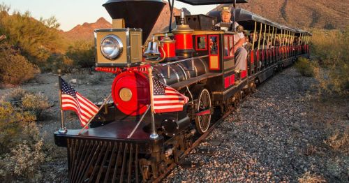 A vintage steam train with a conductor, decorated with American flags, traveling through a desert landscape.