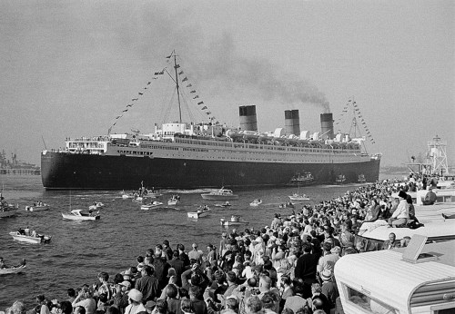 A large ocean liner sails past a crowd gathered along the shore, with small boats in the water and flags flying.