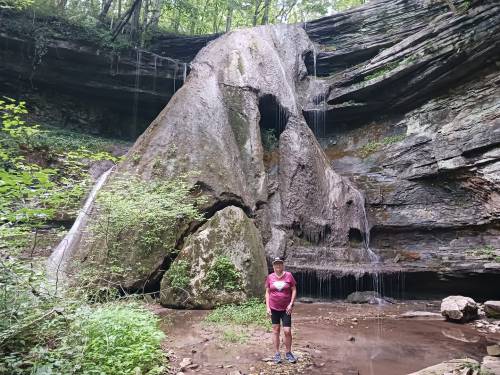 A person stands in front of a large rock formation with water cascading down in a lush, green forest setting.