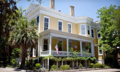 A large yellow Victorian house with a porch, surrounded by greenery and palm trees, under a clear blue sky.