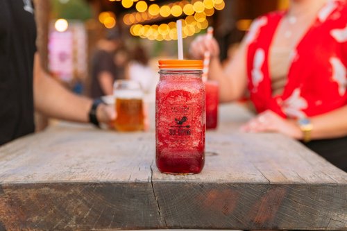 A vibrant red drink in a jar sits on a wooden table, with blurred people and string lights in the background.
