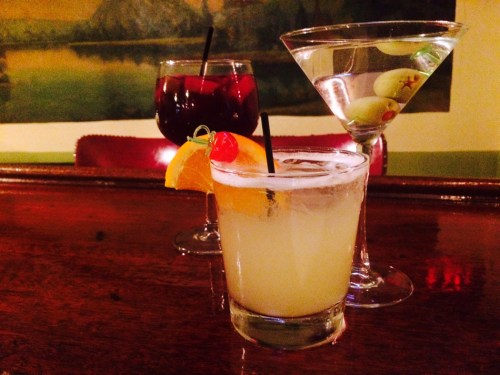 Three cocktails on a wooden bar: a fruity drink with an orange slice, a martini with olives, and a red drink with a cherry.