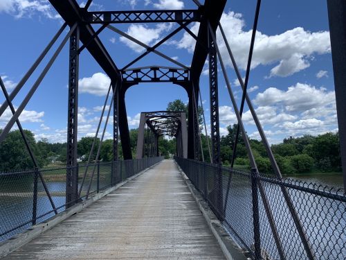 A metal bridge spans a river, with a clear blue sky and fluffy clouds above, surrounded by green trees.