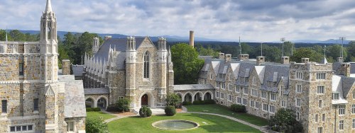 A scenic view of a historic stone college campus with gothic architecture, green lawns, and mountains in the background.