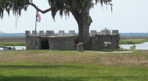 Historic stone fortification with a British flag, surrounded by greenery and water, under a large tree.