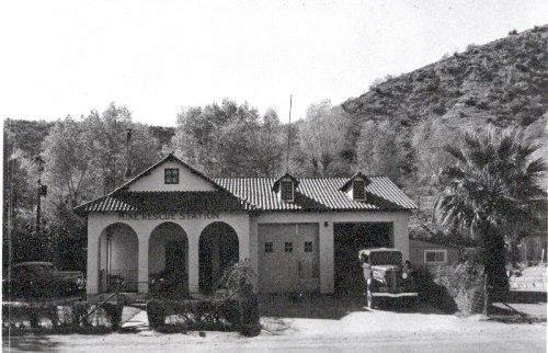 Historic fire rescue station with a tiled roof, arched entrance, and vintage truck parked outside, surrounded by trees.