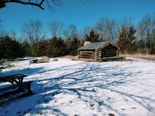 A rustic log cabin surrounded by snow and trees, with a picnic table in the foreground under a clear blue sky.