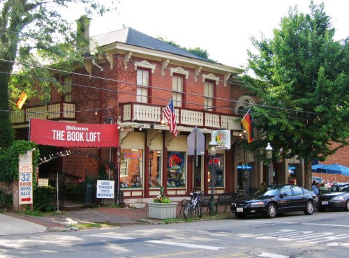 A charming brick building with a sign reading "The Book Loft," featuring flags and outdoor seating.