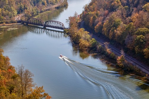 Aerial view of a river with autumn foliage, a boat leaving a wake, and a railway bridge in the background.