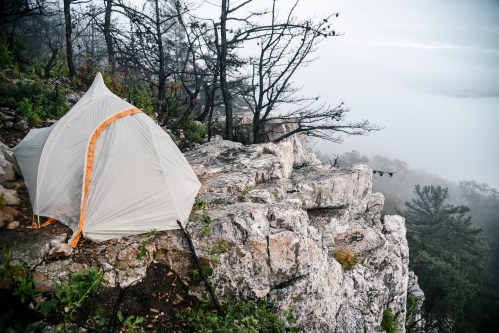 A tent set up on a rocky cliff, surrounded by misty trees and a foggy landscape.