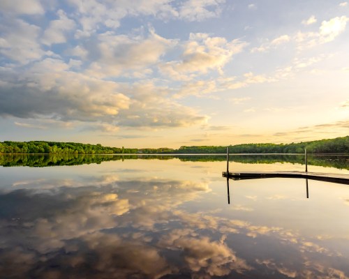 A serene lake at sunset, reflecting clouds and trees, with a wooden dock extending into the water.