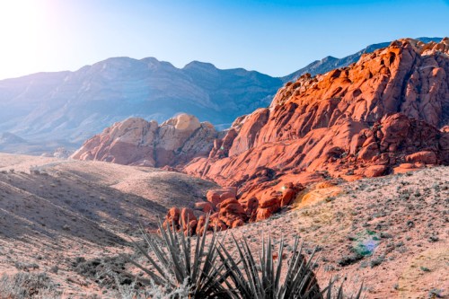 Vibrant red rock formations rise against a clear blue sky, surrounded by desert landscape and distant mountains.