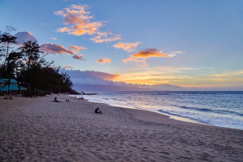 A serene beach at sunset, with soft waves, sandy shore, and silhouettes of people enjoying the view.
