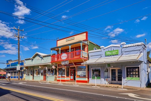 Colorful storefronts line a street under a blue sky with scattered clouds, featuring a mix of shops and a second-story balcony.