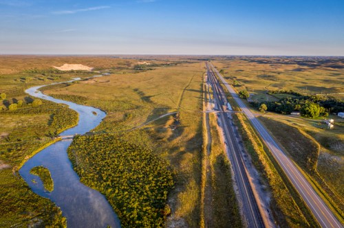 Aerial view of a winding river alongside a highway, surrounded by expansive green fields and rolling hills.