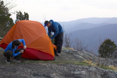 Two campers set up an orange tent on a rocky surface with mountains in the background under a cloudy sky.
