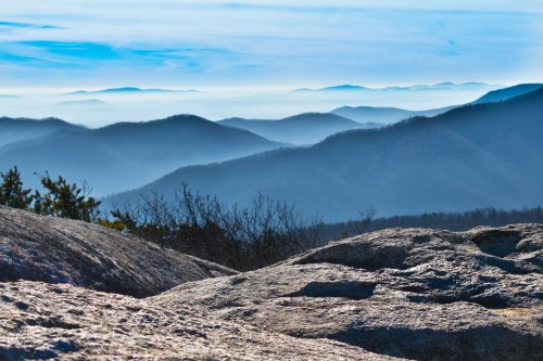 A panoramic view of layered blue mountains under a misty sky, with rocky foreground and distant peaks fading into the horizon.