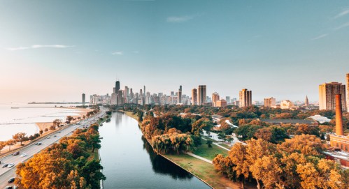 Aerial view of Chicago skyline at sunset, with a river and trees in the foreground, reflecting warm colors.