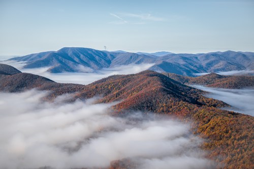 A panoramic view of misty mountains with autumn foliage, showcasing rolling hills and a clear blue sky.