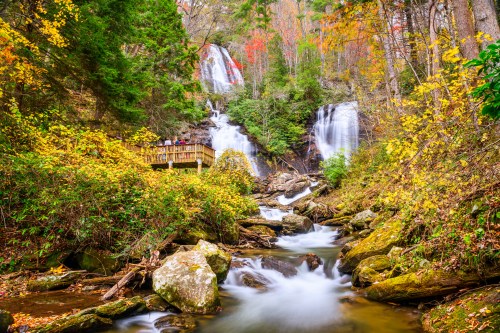 A serene waterfall surrounded by autumn foliage, with a wooden viewing platform and a flowing stream in the foreground.