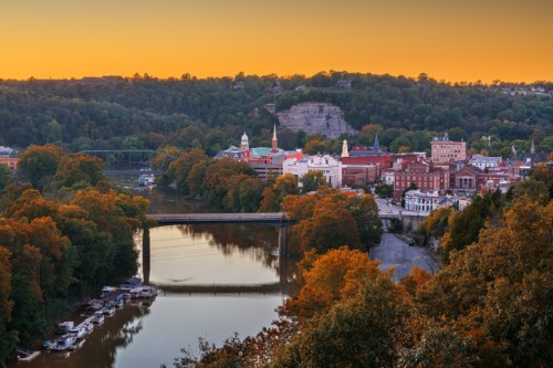 A scenic view of a river town at sunset, with colorful trees, buildings, and a bridge reflecting in the water.