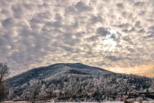 A snow-covered hill under a cloudy sky, with trees glistening in frost and a soft light peeking through the clouds.