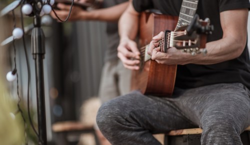 A close-up of a person playing an acoustic guitar, with another musician blurred in the background.