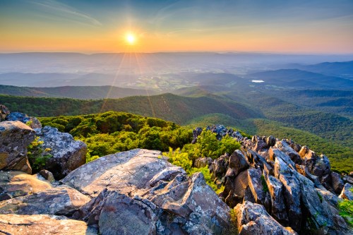 Sunrise over a mountainous landscape, with rocky foreground and lush green valleys stretching into the distance.