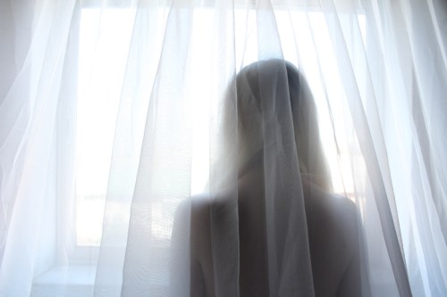 A silhouette of a person standing behind sheer curtains, looking out a window with soft light filtering through.