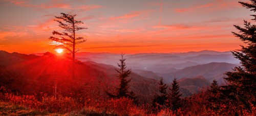 A vibrant sunset over rolling mountains, with silhouetted trees and a warm orange sky.