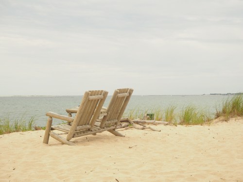 Two wooden lounge chairs on a sandy beach, facing a calm sea under a cloudy sky.