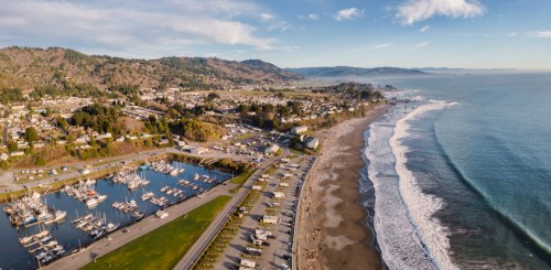 Aerial view of a coastal town with a marina, sandy beach, and rolling waves under a clear blue sky.