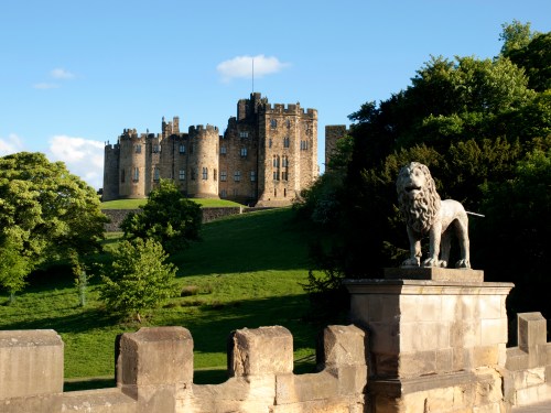 A stone lion statue in the foreground with a historic castle and lush greenery in the background under a clear blue sky.