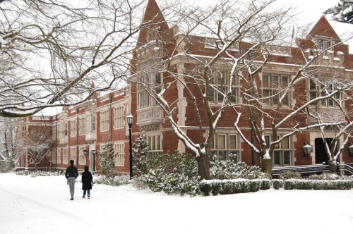 Two people walk along a snow-covered path beside a brick building, surrounded by trees and winter scenery.
