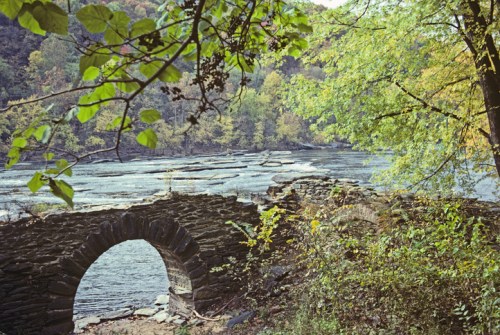 A stone arch bridge over a river, surrounded by colorful autumn foliage and rocky riverbanks.
