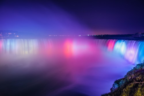 A vibrant night view of a waterfall illuminated with colorful lights, creating a misty, ethereal atmosphere.