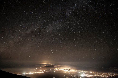 A starry night sky over a coastal city, with twinkling lights and a faint glow on the horizon.