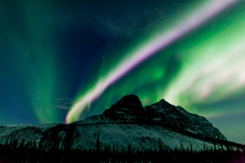 A stunning display of the Northern Lights illuminates the night sky above a snow-covered mountain landscape.