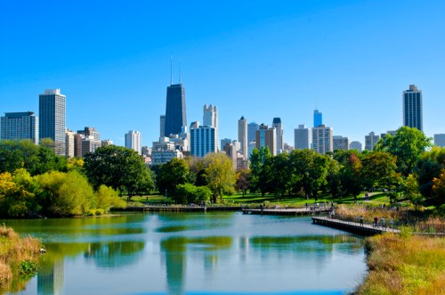 City skyline with tall buildings, green trees, and a calm lake under a clear blue sky.
