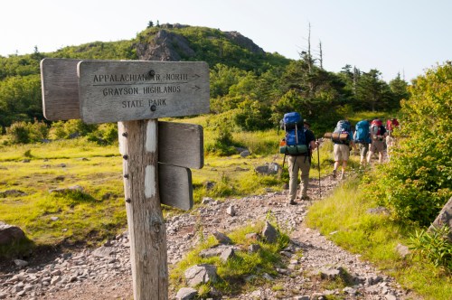 Hikers with backpacks walking on a trail near a wooden sign for the Appalachian Trail in Grayson Highlands State Park.